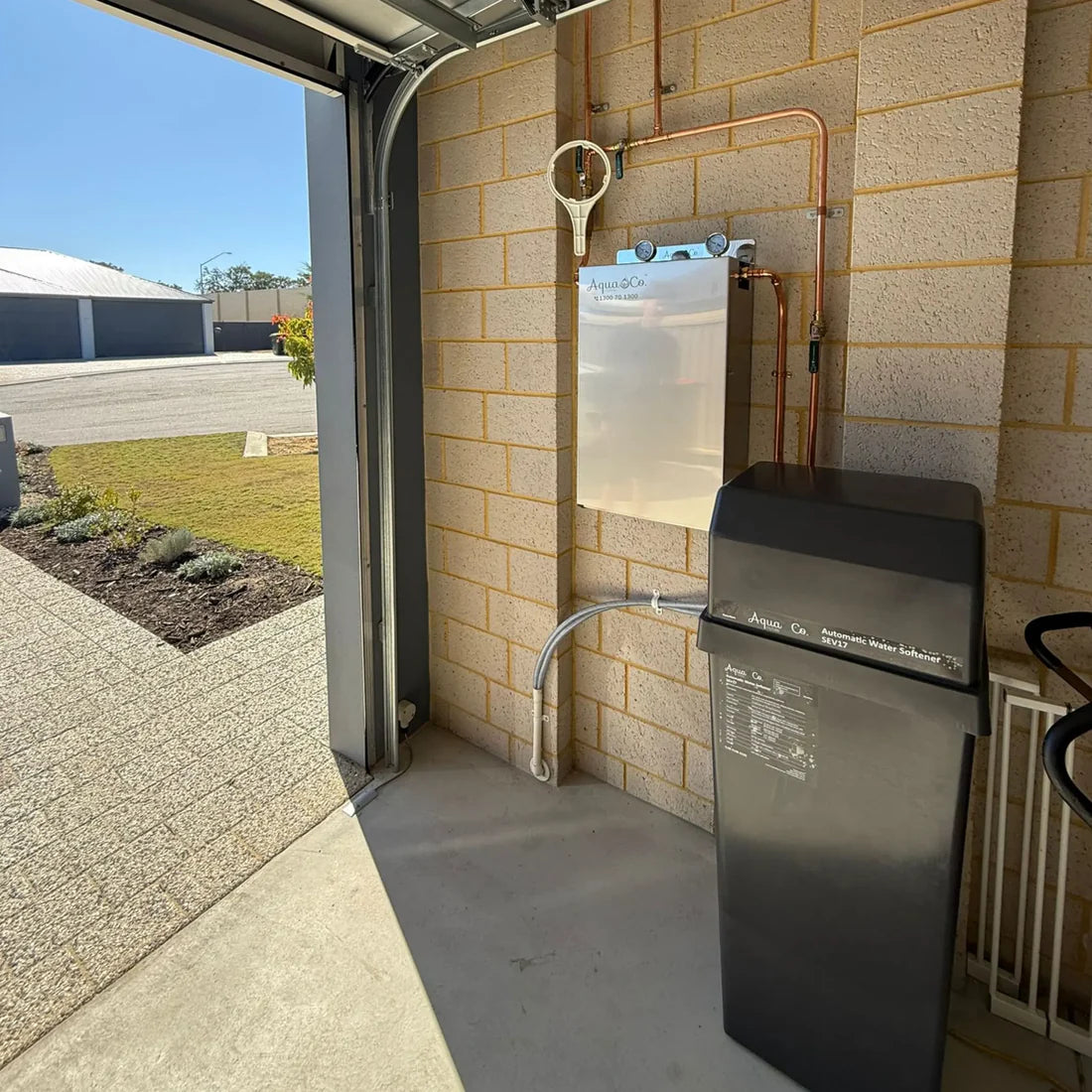 AquaCo water softening filter system installed in a garage with connected copper pipes and outdoor view