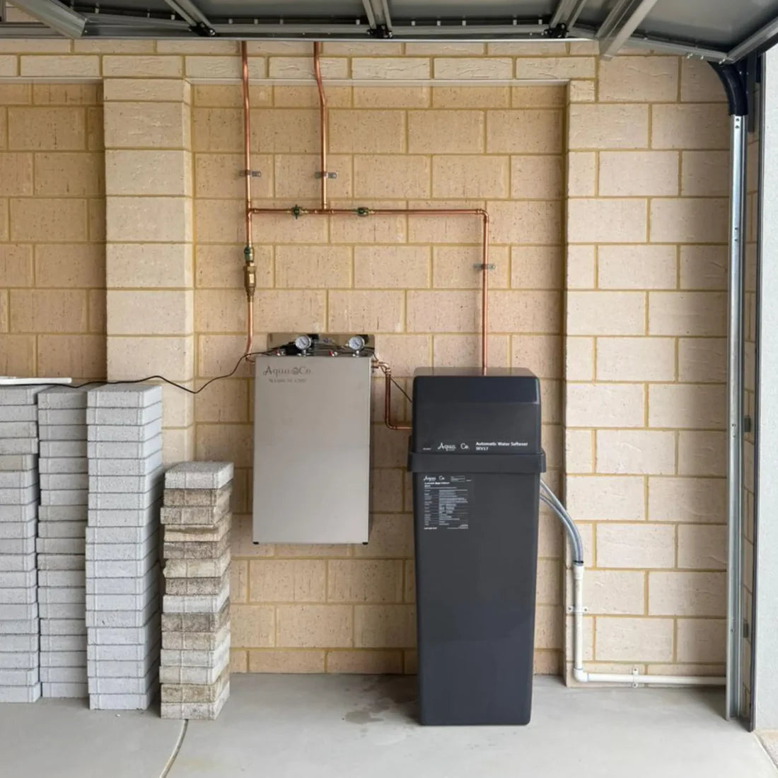 AquaCo water softening filter system installed against beige brick wall with stacked bricks nearby