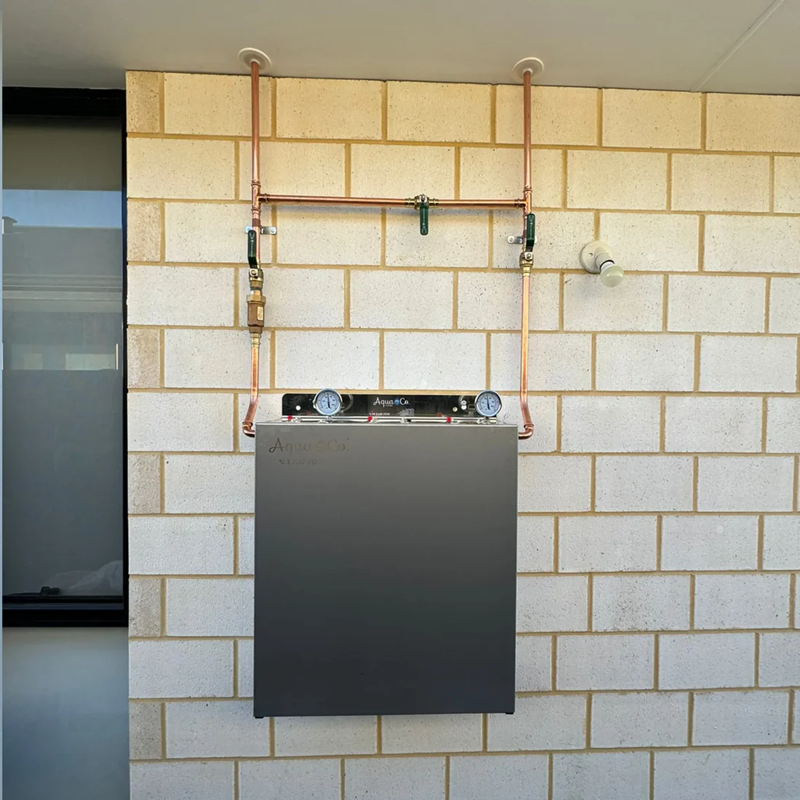 AquaCo whole house water filter system installed on beige brick wall with copper piping and pressure gauges