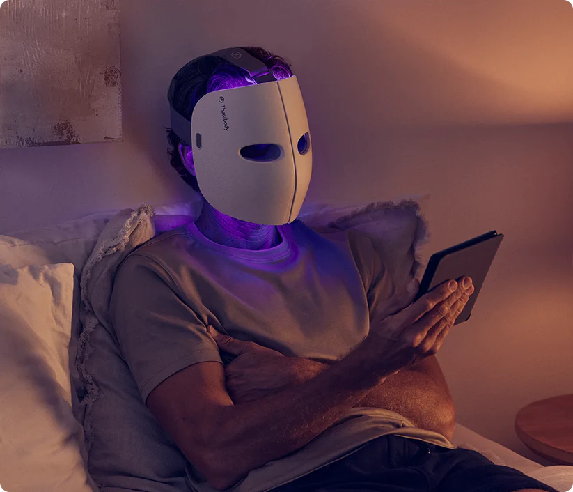 Therabody LED face mask worn by person sitting on bed using tablet for skin therapy