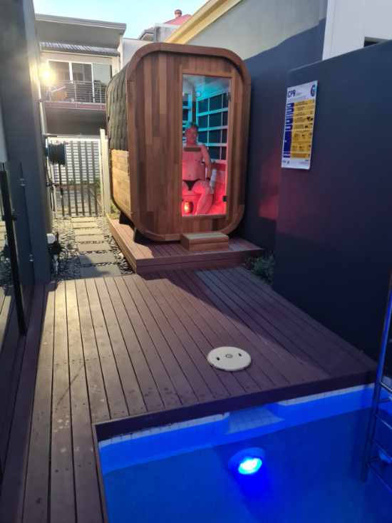 Stryke Recovery outdoor infrared sauna with wooden exterior and glass door next to a pool at dusk
