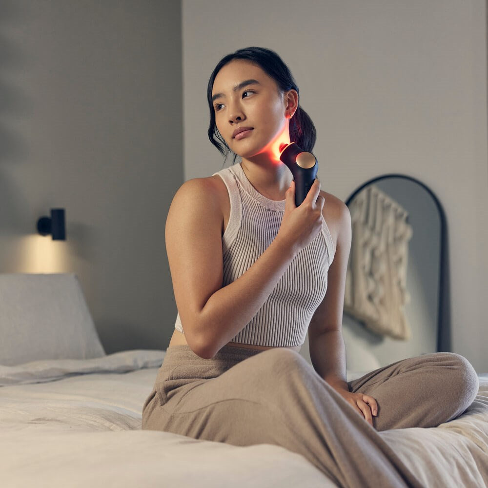 Woman using Therabody theraface pro skincare device with red light therapy on her neck sitting on bed