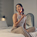 Woman using Therabody theraface pro skincare device with red light therapy on her neck sitting on bed