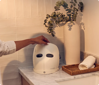 Therabody LED face mask held by hand on bathroom counter with towel and vase decor