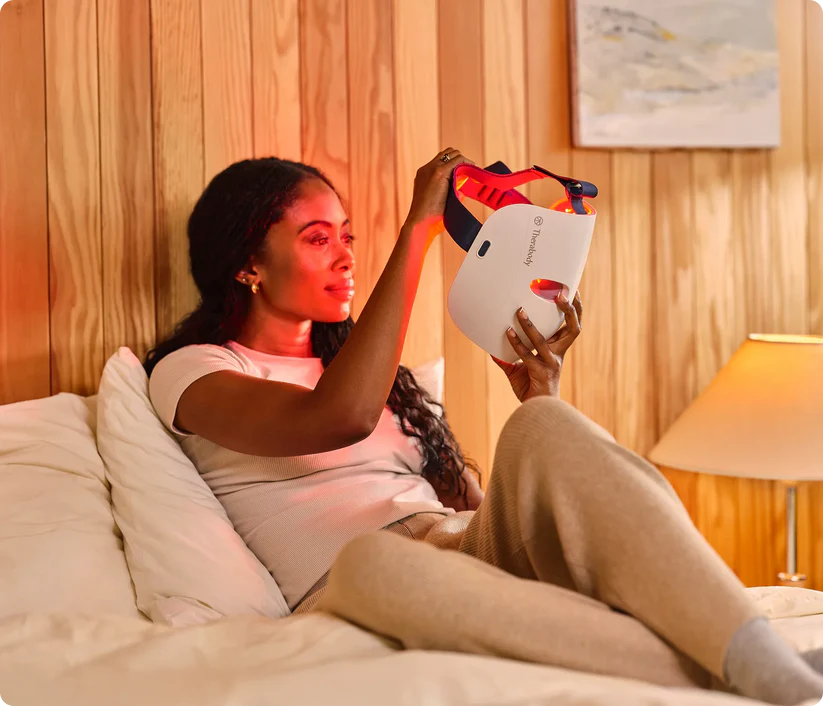 Woman sitting on bed holding Therabody LED mask with red light therapy for skincare
