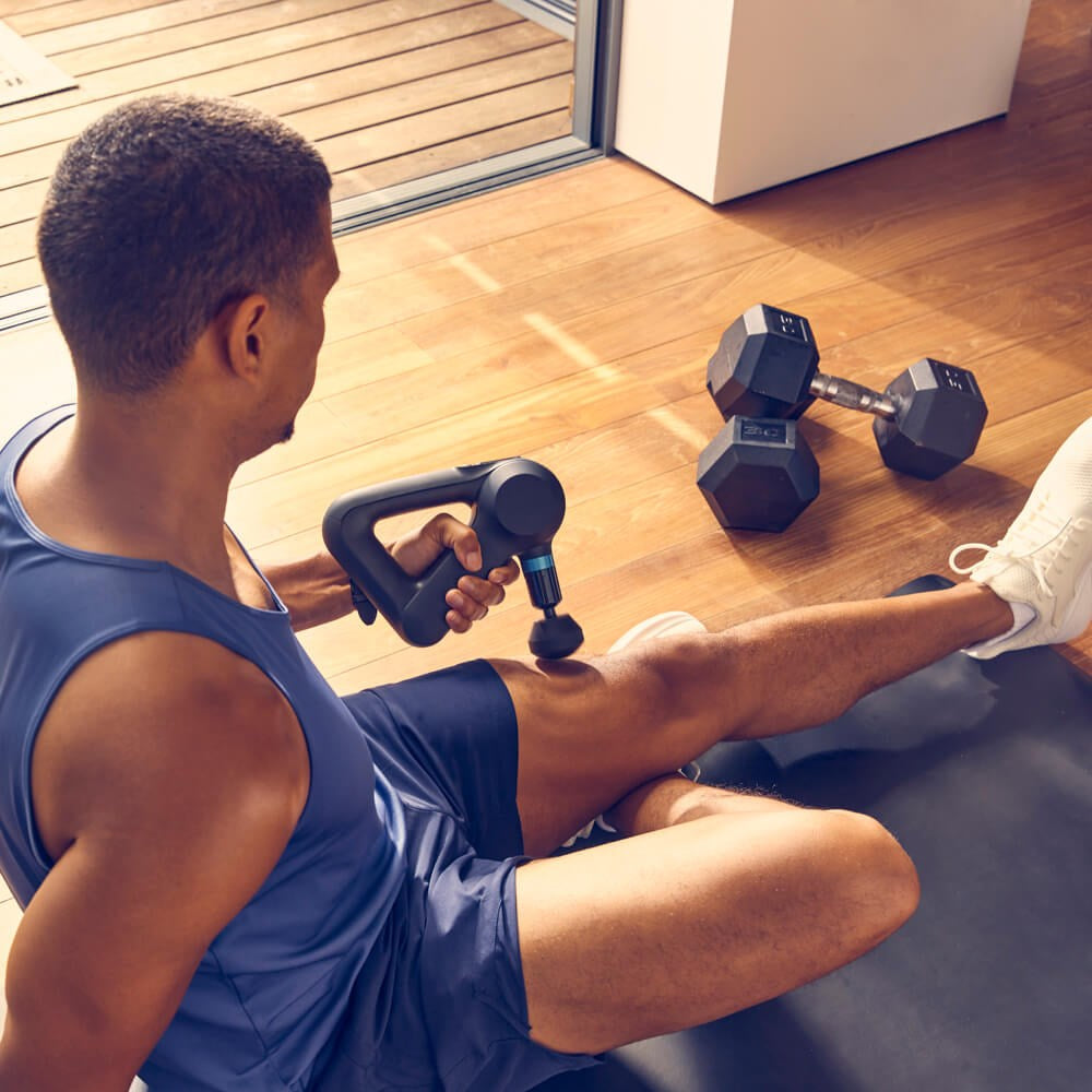 Man using Therabody massage gun on leg muscles while sitting on exercise mat near dumbbells