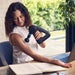 Woman using Therabody massage gun on arm while sitting at desk with laptop and open book